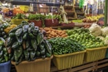 A vibrant market stall displays a variety of fresh vegetables, including green peppers, limes, oranges, cauliflower, and leafy greens. The stall is neatly organized with the produce placed in colorful baskets and bins, creating a lively and inviting scene.
