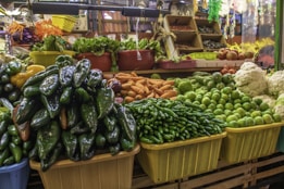 A vibrant market stall displays a variety of fresh vegetables, including green peppers, limes, oranges, cauliflower, and leafy greens. The stall is neatly organized with the produce placed in colorful baskets and bins, creating a lively and inviting scene.