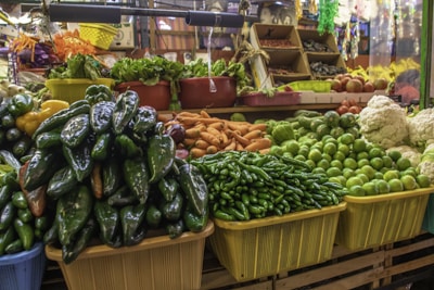 A vibrant market stall displays a variety of fresh vegetables, including green peppers, limes, oranges, cauliflower, and leafy greens. The stall is neatly organized with the produce placed in colorful baskets and bins, creating a lively and inviting scene.