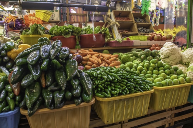 A vibrant market stall displays a variety of fresh vegetables, including green peppers, limes, oranges, cauliflower, and leafy greens. The stall is neatly organized with the produce placed in colorful baskets and bins, creating a lively and inviting scene.