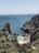 A secluded beach nestled between rocky cliffs with clear blue water. People are enjoying the sun on the sand, swimming, and exploring the natural surroundings. The landscape is lush with greenery covering the rugged cliffs.