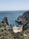A secluded beach nestled between rocky cliffs with clear blue water. People are enjoying the sun on the sand, swimming, and exploring the natural surroundings. The landscape is lush with greenery covering the rugged cliffs.