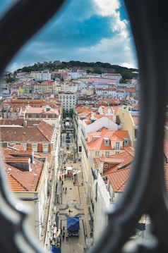A picturesque urban view showcasing a narrow street lined with colorful historic buildings featuring red-tiled roofs. Crowds of people can be seen walking along the street, with some gathered under a blue canopy in a small square. The perspective is framed by the blurred edges of a window or an architectural structure, enhancing the depth of the image. In the background, a lush green hilltop with a castle overlooks the cityscape under a partly cloudy sky.