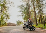 A couple embracing near a vintage motorcycle parked by a palm tree-lined coastal road under soft Mediterranean light.