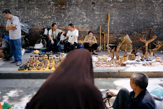 Several people are sitting on the sidewalk, engaging in conversation near a display of small pottery items and intricately carved wooden figures. The backdrop is a textured brick wall, adding an urban feel to the scene. The ground is partially shaded by the trees, which scatter patches of sunlight.