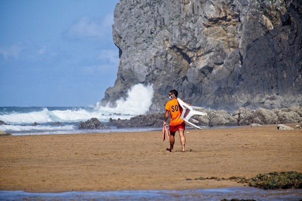 A lifeguard in an orange SOS shirt is walking along a sandy beach. He is carrying a folded white plastic chair over his shoulder and holding a rescue buoy. Waves crash against rocky cliffs in the background under a partly cloudy sky.