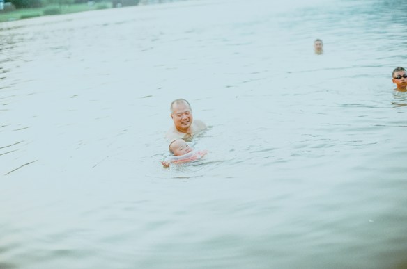 Several people are swimming in a body of water, with a man holding a baby in a flotation device. The scene is serene and captures a family-friendly atmosphere.