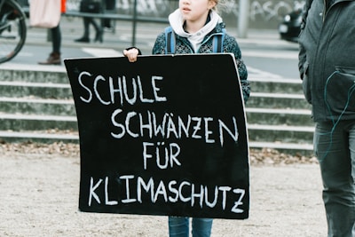 A person is holding a black protest sign with white text that reads 'Schule schwänzen für Klimaschutz' which translates to 'Skipping school for climate protection'. The surroundings indicate an outdoor public space, possibly during a demonstration or protest.