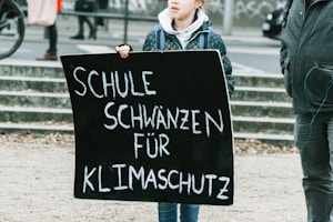 A person is holding a black protest sign with white text that reads 'Schule schwänzen für Klimaschutz' which translates to 'Skipping school for climate protection'. The surroundings indicate an outdoor public space, possibly during a demonstration or protest.