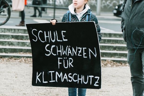 A person is holding a black protest sign with white text that reads 'Schule schw&auml;nzen f&uuml;r Klimaschutz' which translates to 'Skipping school for climate protection'. The surroundings indicate an outdoor public space, possibly during a demonstration or protest.