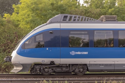 A modern blue and white train travels along a railway track, surrounded by lush green vegetation. The design features a streamlined front and large windows with passengers visible inside.