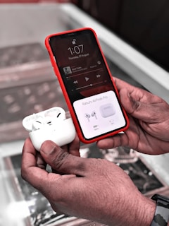 A person holds a smartphone with a red case showing a music player interface and AirPods Pro pairing screen. In the other hand, there is a pair of AirPods Pro in its open charging case. The background appears blurred, focusing on the hands and the devices.