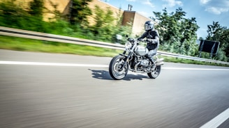 A motorcyclist wearing protective gear and a helmet rides a classic bike down a highway. The background is blurred, suggesting high speed, with a cloudy sky above and greenery beside the road.