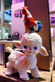 A cute ceramic lamb with blue eyes and a textured wool-like surface stands on a wooden shelf. On top of the lamb is a colorful toy rooster, painted in vibrant red, blue, and white colors. The background shows a blurred indoor setting with soft lighting and other objects slightly out of focus.