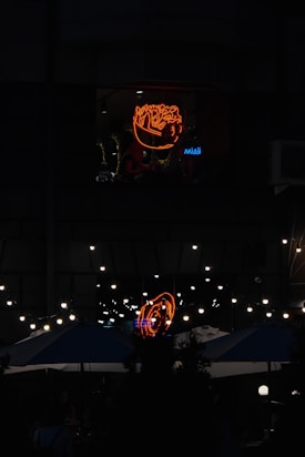 Neon lights featuring a brightly lit design of a ramen bowl are prominent on a dark facade. Below, string lights create a warm ambiance over outdoor seating areas equipped with large umbrellas. The scene suggests a nighttime urban environment with elements of street or restaurant culture.