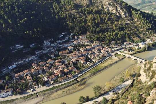A small town with traditional red-roofed houses nestled at the base of a forested hillside. A river flows alongside the town, crossed by an arched stone bridge. The town is surrounded by lush greenery and rocky terrain.
