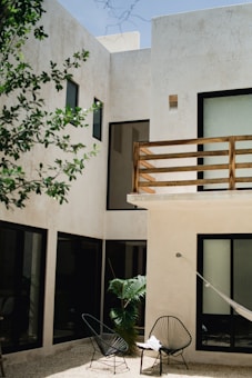 A modern courtyard with a minimalist design featuring large beige walls and several black-framed windows. In the foreground, two wire chairs are placed on a gravel surface next to a green potted plant. A hammock is tied beside a wooden balcony railing on the upper level.
