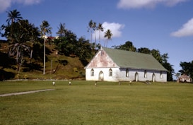 A stone church with a green roof is set against a hill with several palm trees. The church is surrounded by a grassy field, where a few children are playing. The sky is clear with some scattered clouds.