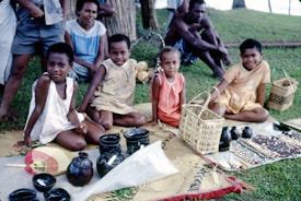 A group of children is seated on woven mats on the grass with handcrafted items such as baskets, pottery, and necklaces displayed in front of them. Two adults sit behind, observing. The setting appears outdoors, with a tree and more greenery in the background.