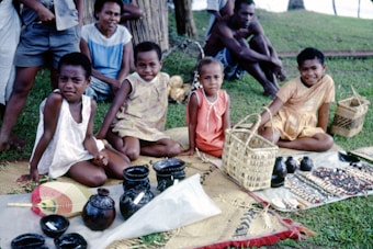 A group of children is seated on woven mats on the grass with handcrafted items such as baskets, pottery, and necklaces displayed in front of them. Two adults sit behind, observing. The setting appears outdoors, with a tree and more greenery in the background.