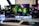 A gaming setup featuring a central monitor with a vibrant display showing 'The Spirit of Gaming'. On either side, there are gaming PCs with illuminated components and transparent cases. Headsets hang from the side of the monitor. The desk also has a black keyboard and mouse, and a branded mousepad.