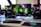 A gaming setup featuring a central monitor with a vibrant display showing 'The Spirit of Gaming'. On either side, there are gaming PCs with illuminated components and transparent cases. Headsets hang from the side of the monitor. The desk also has a black keyboard and mouse, and a branded mousepad.