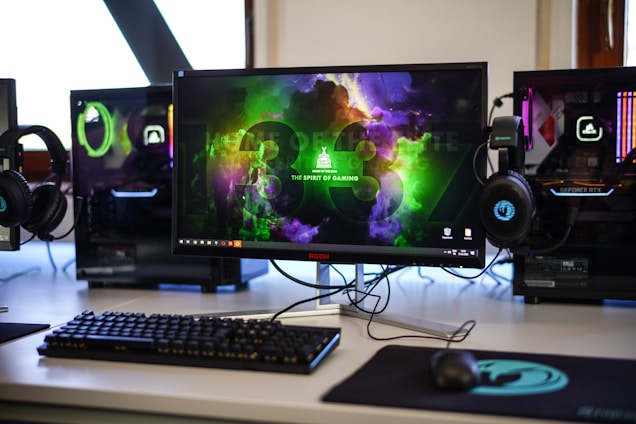 A gaming setup featuring a central monitor with a vibrant display showing 'The Spirit of Gaming'. On either side, there are gaming PCs with illuminated components and transparent cases. Headsets hang from the side of the monitor. The desk also has a black keyboard and mouse, and a branded mousepad.