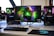 A gaming setup featuring a central monitor with a vibrant display showing 'The Spirit of Gaming'. On either side, there are gaming PCs with illuminated components and transparent cases. Headsets hang from the side of the monitor. The desk also has a black keyboard and mouse, and a branded mousepad.