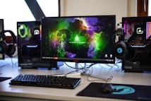 A gaming setup featuring a central monitor with a vibrant display showing 'The Spirit of Gaming'. On either side, there are gaming PCs with illuminated components and transparent cases. Headsets hang from the side of the monitor. The desk also has a black keyboard and mouse, and a branded mousepad.