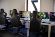 A row of gaming stations equipped with large monitors, gaming chairs, keyboards, and headphones. The setup is organized and ready for gaming, featuring a colorful screen display. The room has a modern and tech-savvy ambiance.