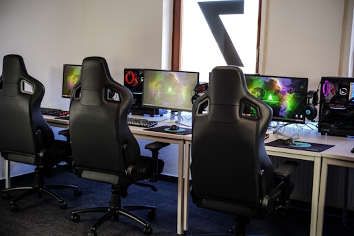 A row of gaming stations equipped with large monitors, gaming chairs, keyboards, and headphones. The setup is organized and ready for gaming, featuring a colorful screen display. The room has a modern and tech-savvy ambiance.