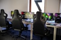 A row of gaming stations equipped with large monitors, gaming chairs, keyboards, and headphones. The setup is organized and ready for gaming, featuring a colorful screen display. The room has a modern and tech-savvy ambiance.