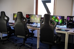 A row of gaming stations equipped with large monitors, gaming chairs, keyboards, and headphones. The setup is organized and ready for gaming, featuring a colorful screen display. The room has a modern and tech-savvy ambiance.