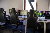 A row of gaming stations equipped with large monitors, gaming chairs, keyboards, and headphones. The setup is organized and ready for gaming, featuring a colorful screen display. The room has a modern and tech-savvy ambiance.