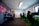 A well-lit gaming room features multiple gaming stations with high-backed chairs and monitors. The room has a minimalist setup with green and black color tones. Large posters adorn the walls, and the room appears clean and organized.