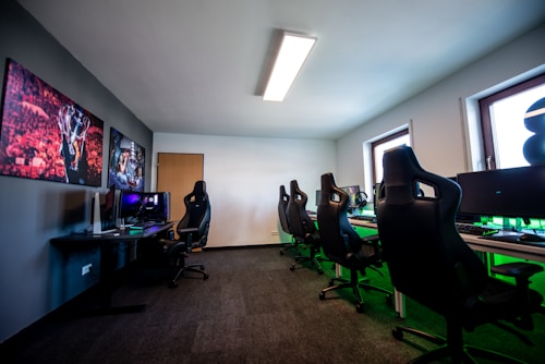 A well-lit gaming room features multiple gaming stations with high-backed chairs and monitors. The room has a minimalist setup with green and black color tones. Large posters adorn the walls, and the room appears clean and organized.