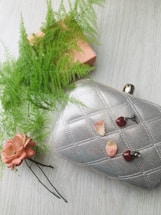 A silver quilted clutch purse with metallic sheen placed on a light-colored wooden surface. It is adorned with two pink rose petals and a pair of heart-shaped red earrings. A delicate pink flower and light green ferns are arranged alongside, providing a fresh and elegant display.