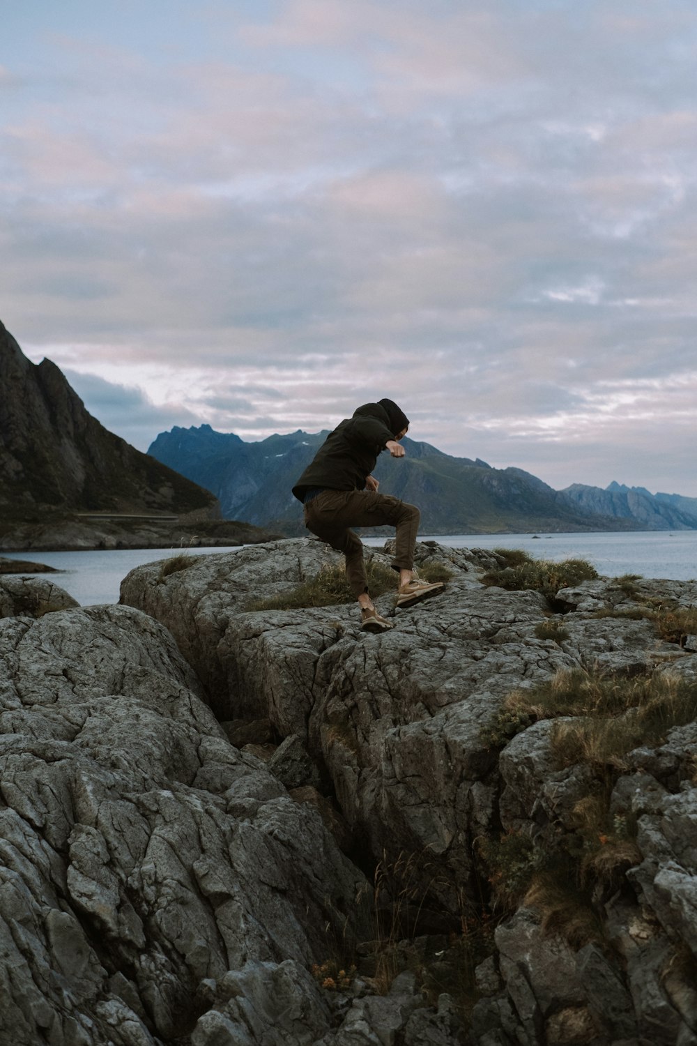 man in black t-shirt and gray pants sitting on rock formation near body of water