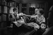 In a shop with shelves stacked with chocolate bars, a woman is interacting with a salesperson behind the counter. She appears to be making a purchase, holding an item in one hand and gesturing with the other. A man and a young boy stand beside her, observing the transaction.