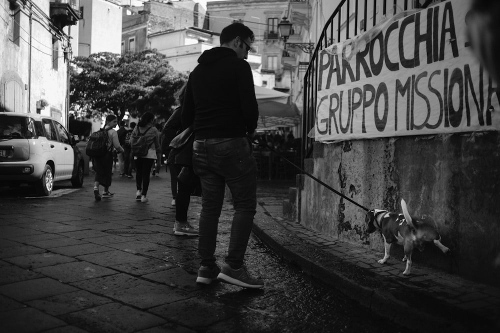 man in black jacket and blue denim jeans walking on sidewalk with dog