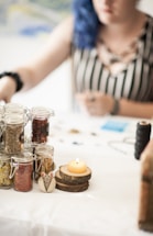 A table is set with several glass jars containing herbs or spices, a lit candle on a wooden coaster, and some crafting materials in the background. A person is partially visible, sitting at the table. The setting appears relaxed and artistic.
