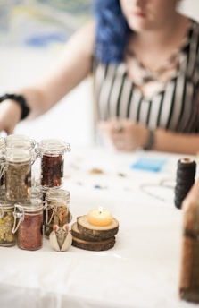 A table is set with several glass jars containing herbs or spices, a lit candle on a wooden coaster, and some crafting materials in the background. A person is partially visible, sitting at the table. The setting appears relaxed and artistic.