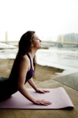 A woman is practicing yoga on a pink mat in an outdoor setting by a river with a bridge and cityscape in the background. She is in an upward-facing position, with her eyes closed, conveying a sense of calm and focus.