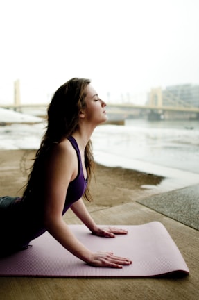 A woman is practicing yoga on a pink mat in an outdoor setting by a river with a bridge and cityscape in the background. She is in an upward-facing position, with her eyes closed, conveying a sense of calm and focus.