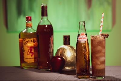 Several bottles of beverages, including a whiskey bottle labeled Fireball, a Mundet apple soda, and a golden spherical bottle, are arranged on a table. There is a glass filled with ice and a straw, and an apple is placed next to the bottles. The background features green hues, creating a warm and inviting atmosphere.