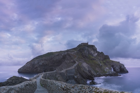 A rugged island connected by a winding stone walkway, with a building perched atop the hill. The scene is enveloped in a serene atmosphere, with soft clouds filling the sky and calm ocean waters surrounding the island. The natural rock formations and lush greenery add to the dramatic landscape.