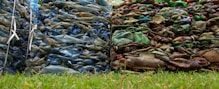 Piles of compacted plastic bottles and containers, with one stack appearing blue and the other brown and green, are placed side by side on a grassy surface.