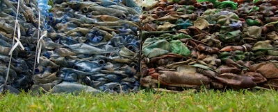 Piles of compacted plastic bottles and containers, with one stack appearing blue and the other brown and green, are placed side by side on a grassy surface.