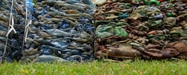 Piles of compacted plastic bottles and containers, with one stack appearing blue and the other brown and green, are placed side by side on a grassy surface.