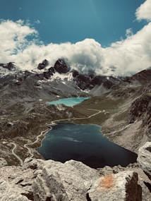A breathtaking mountainous landscape with rugged peaks partially covered in snow and clouds looming above. Two serene lakes, one near the bottom of the image and another further back, exhibit deep blue and turquoise hues. A winding road snakes through the rocky terrain, offering a sense of scale and depth.